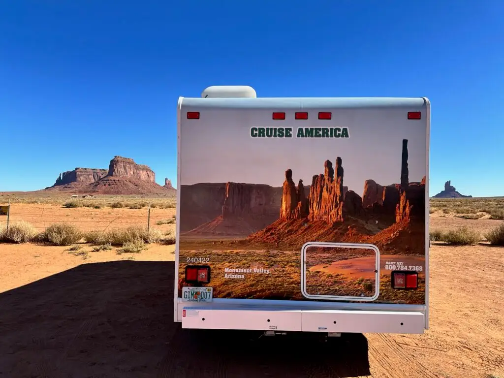 Back of a Cruise America rental RV with back side photo of red rock spires at Monument Valley in Arizona, parked facing actual red rock spires at Monument Valley in Arizona.