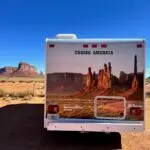 Back of a Cruise America rental RV with back side photo of red rock spires at Monument Valley in Arizona, parked facing actual red rock spires at Monument Valley in Arizona.