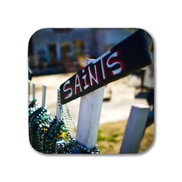 A coaster featuring a New Orleans picture of a white picket fence decorated with Mardi Gras beads with a sign saying, "Saints" nailed to the top