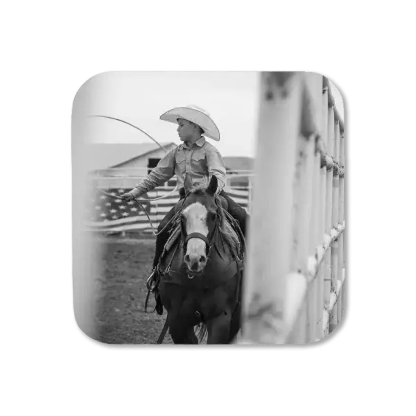 A boy spins his lasso while on horseback next to a fence, with American flag in background